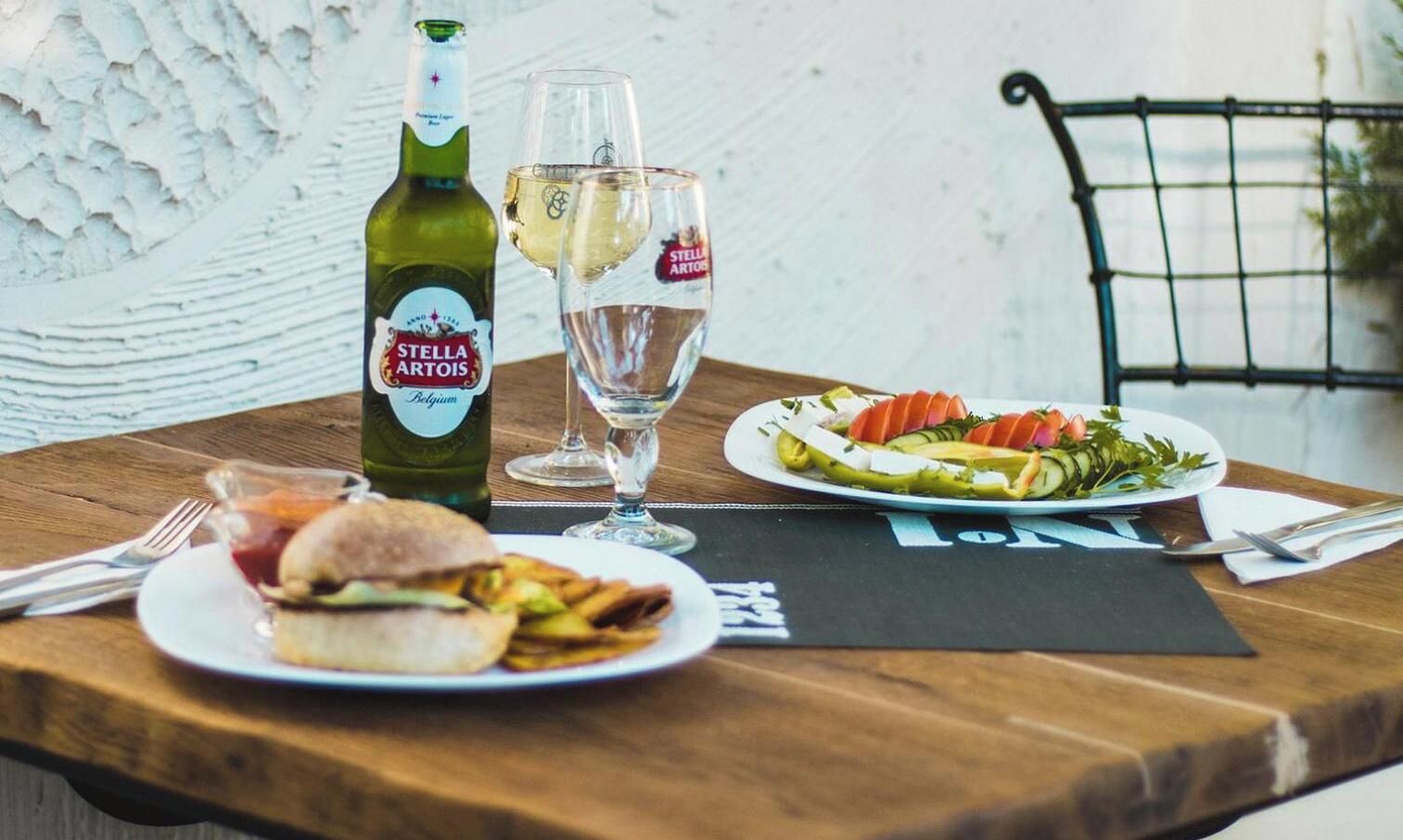 A table with a bottle of Stella Artois, a glass of white wine, a sandwich, a plate of salad, and cutlery.