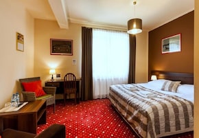Una habitación de hotel con cama, escritorio, silla, lámpara y ventana con cortinas.