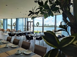 A dining area with tables set for service, large windows offering a view of a body of water, and indoor plants. - Domein Polderwind (Zuienkerke)