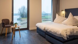 A bedroom with a bed, a chair, a small table, and a view of a body of water through a window. - Domein Polderwind (Zuienkerke)
