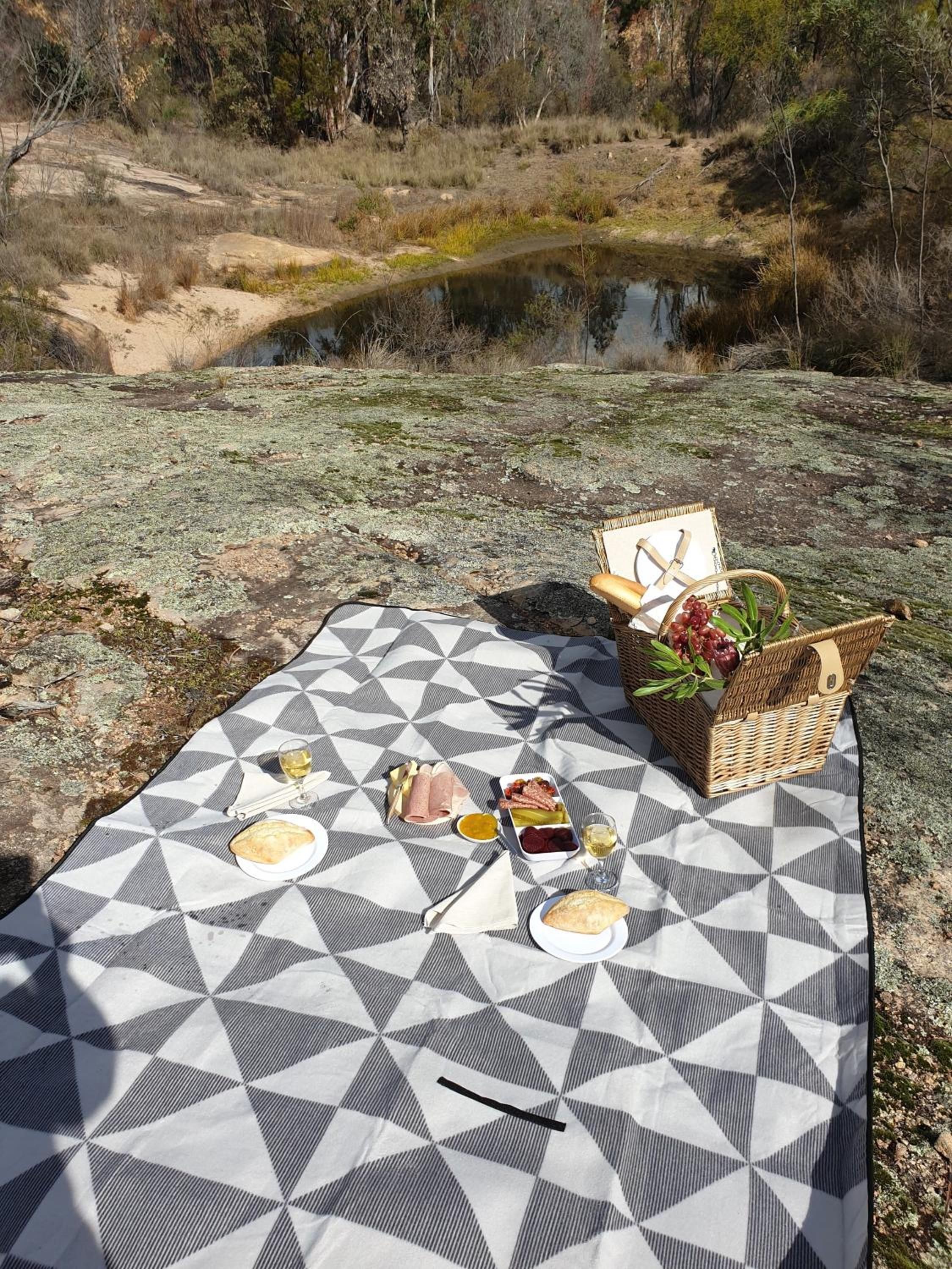 A picnic setup with a blanket, food, and a basket on a rocky terrain near a body of water.