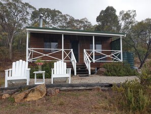A wooden cabin with a porch, two white chairs, a small table, and a water tank in the background.