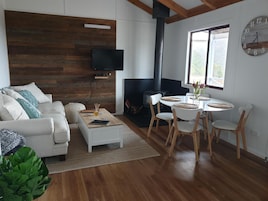 A modern living room with a white sofa, a coffee table, a fireplace, a dining area with chairs, and a wall-mounted TV.