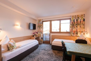 A hotel room with two single beds, a small table, a TV mounted on the wall, and a window with curtains.