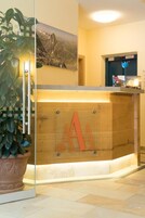 Un mostrador de recepción con panel de madera y una gran letra 'A', una planta en maceta a la izquierda y un cuadro enmarcado sobre el mostrador.