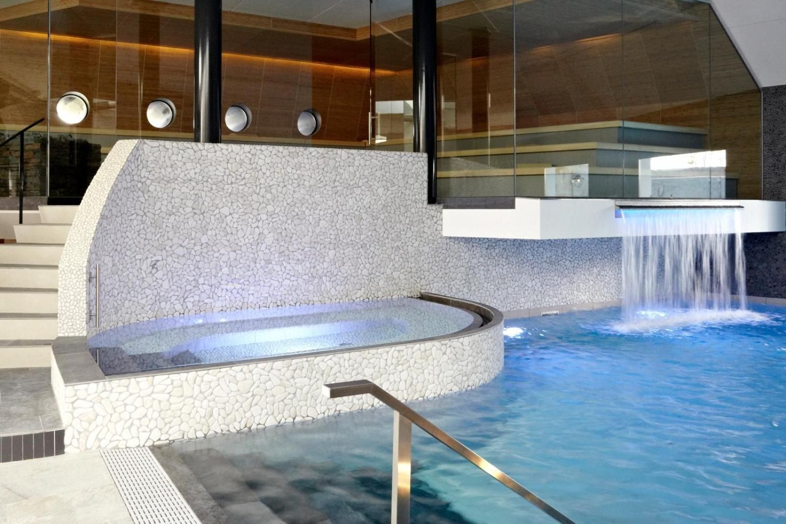 A modern indoor pool area with a hot tub, a waterfall feature, and a stone wall.