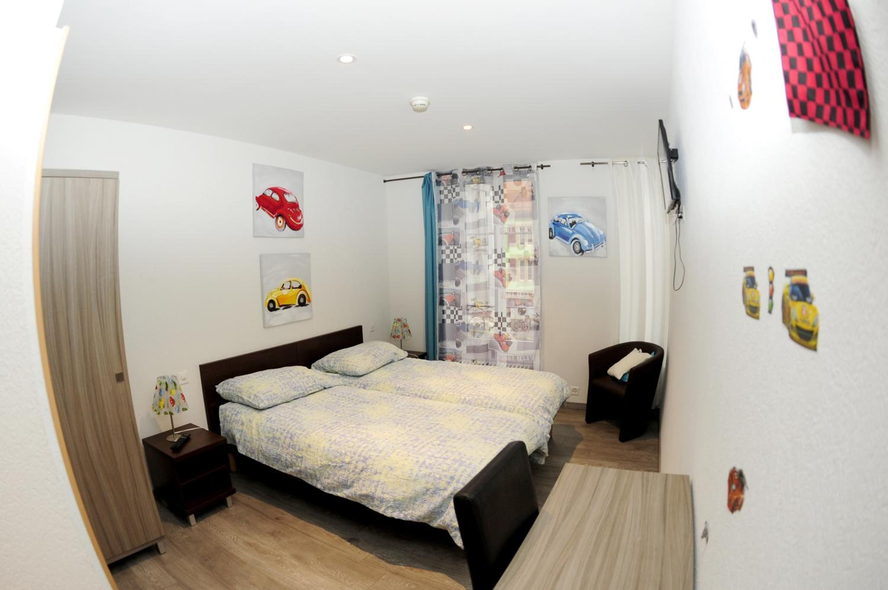Quarto com cama, mesas de cabeceira, um criado-mudo, TV fixada na parede e cortina estampada com tema automotivo.