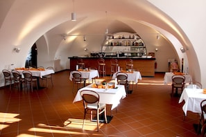 A restaurant with multiple tables and chairs, a bar area with shelves of bottles, and a tiled floor.