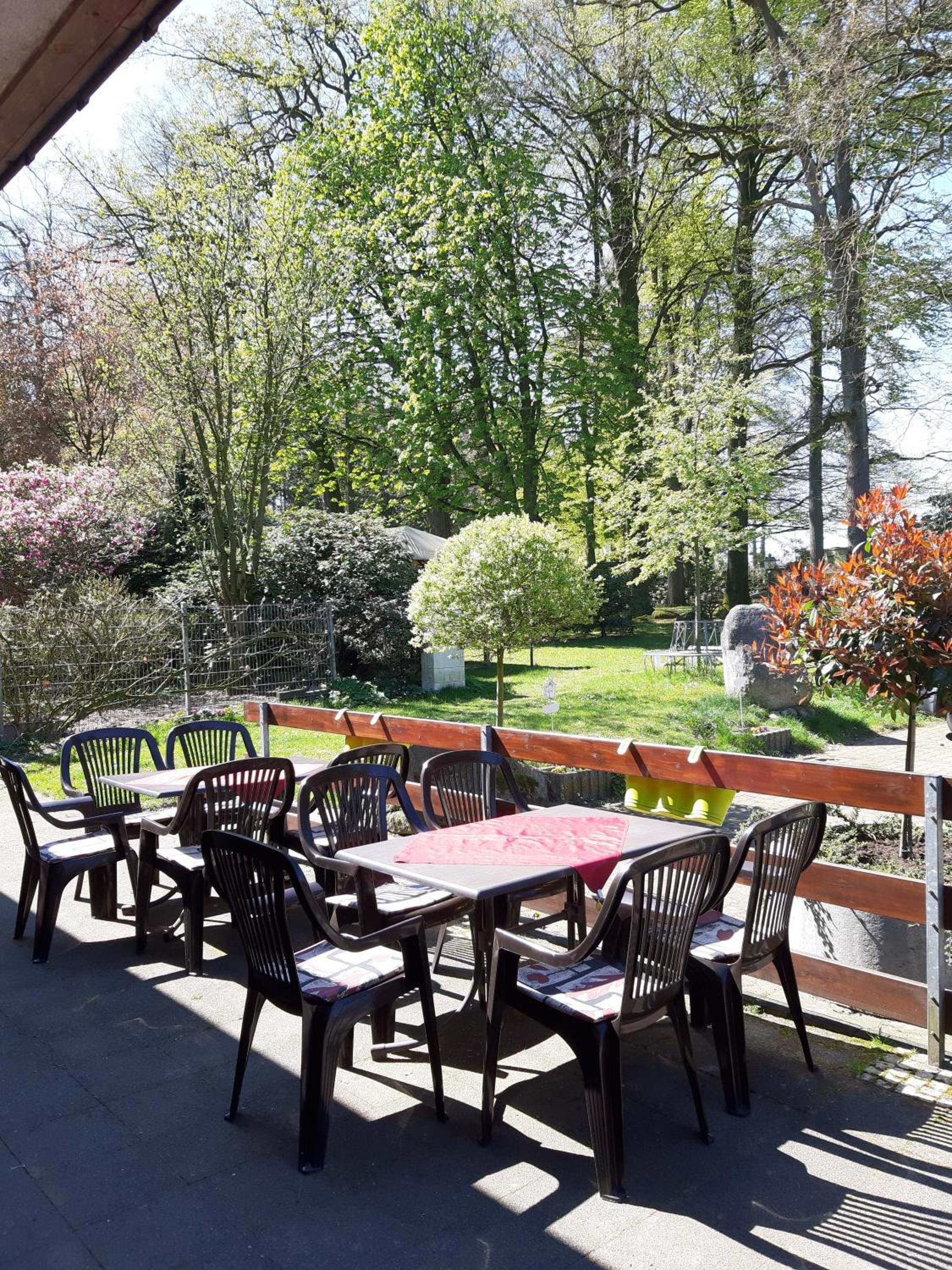 An outdoor seating area with tables and chairs, surrounded by trees and a stone wall.