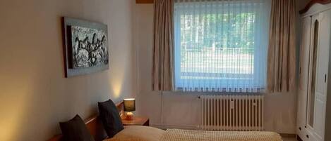 Un dormitorio con techo de madera, una cama con dos almohadas, una mesita de noche con lámpara, una ventana con cortinas y un radiador.