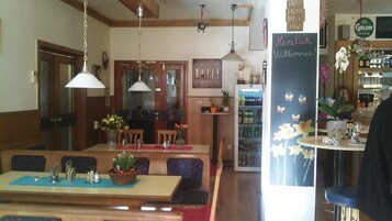 Interior de un restaurante con mobiliario de madera, lámparas colgantes y una pizarra con un mensaje de bienvenida.