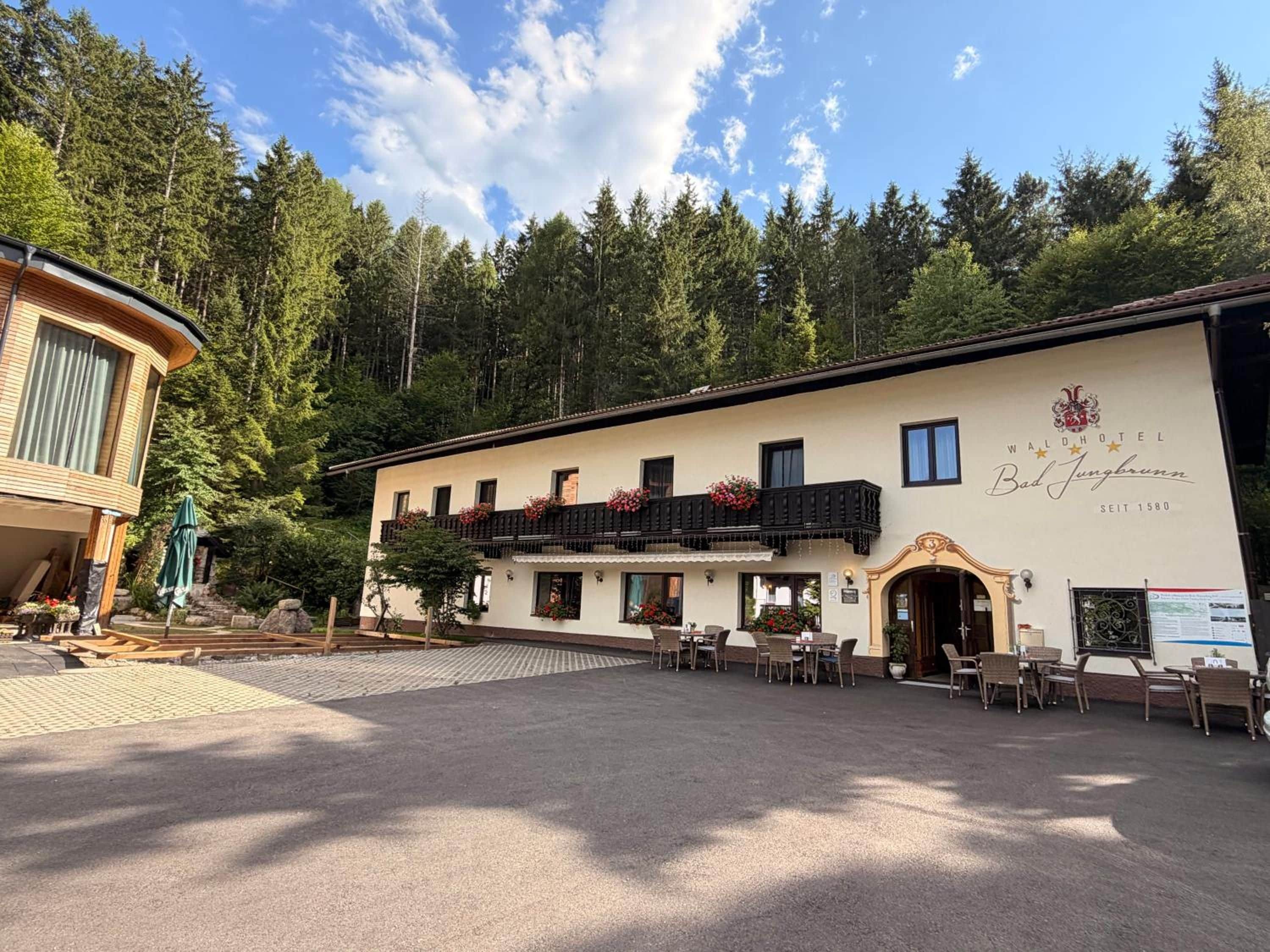 Un hotel con balcón, mobiliario exterior y un paisaje boscoso de fondo.
