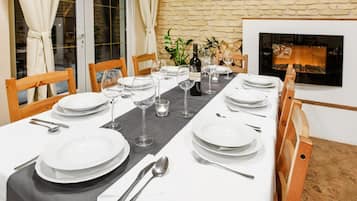 Mesa de jantar posta para quatro, com pratos brancos, talheres e copos de vinho, em um quarto com parede de pedra e lareira.