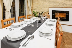 Mesa de jantar posta para quatro, com pratos brancos, talheres e copos de vinho, em um quarto com parede de pedra e lareira.