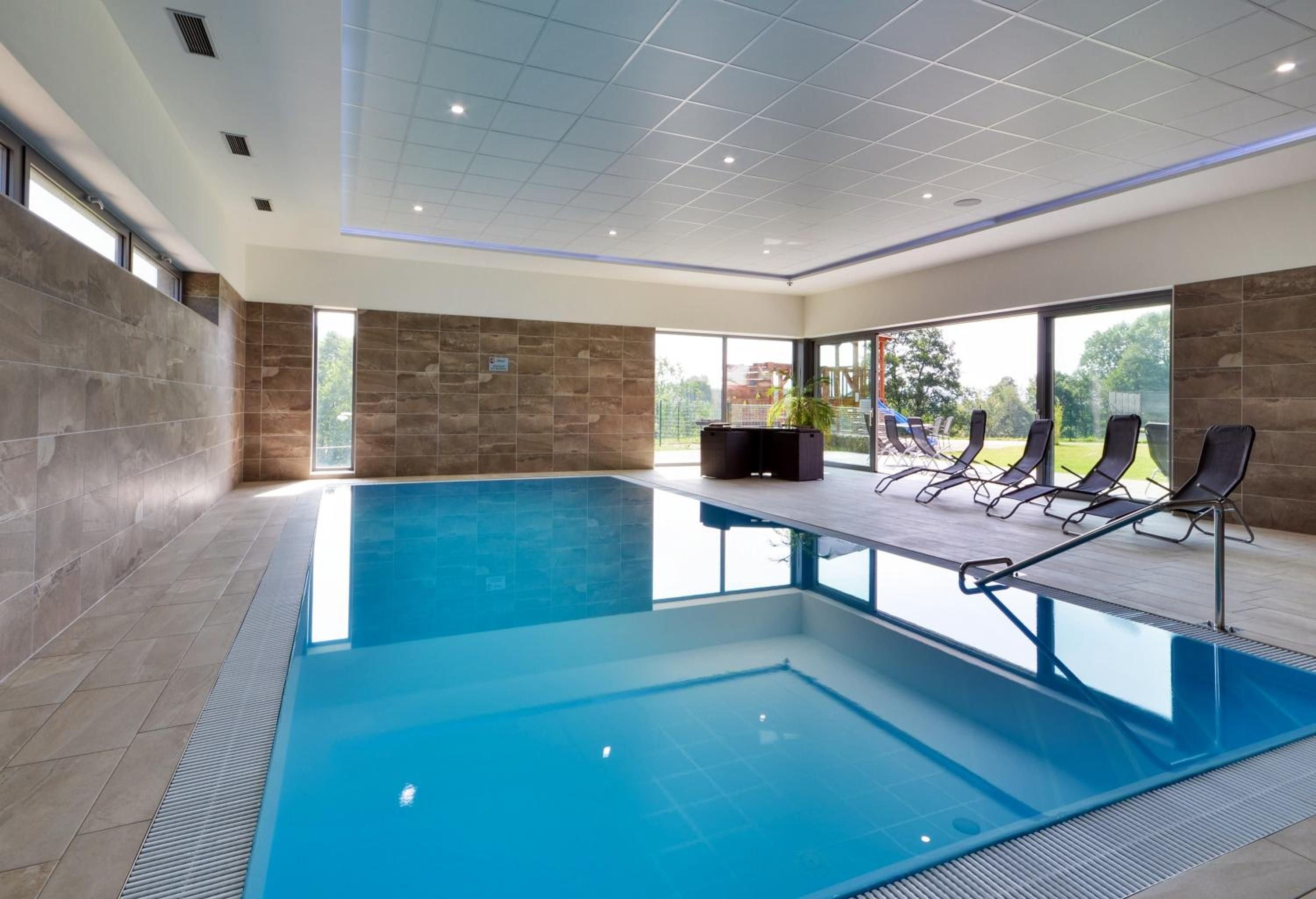 A modern indoor swimming pool with clear blue water, surrounded by lounge chairs and a stone wall.