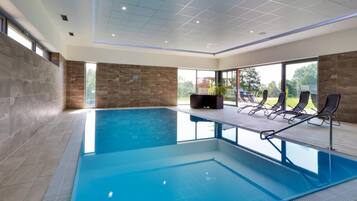 A modern indoor swimming pool with clear blue water, surrounded by lounge chairs and a stone wall.