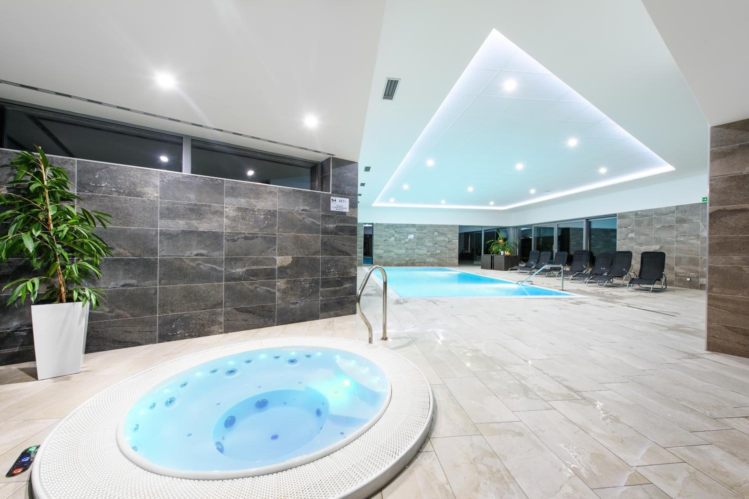 A modern indoor swimming pool area with a hot tub, lounge chairs, and a potted plant.