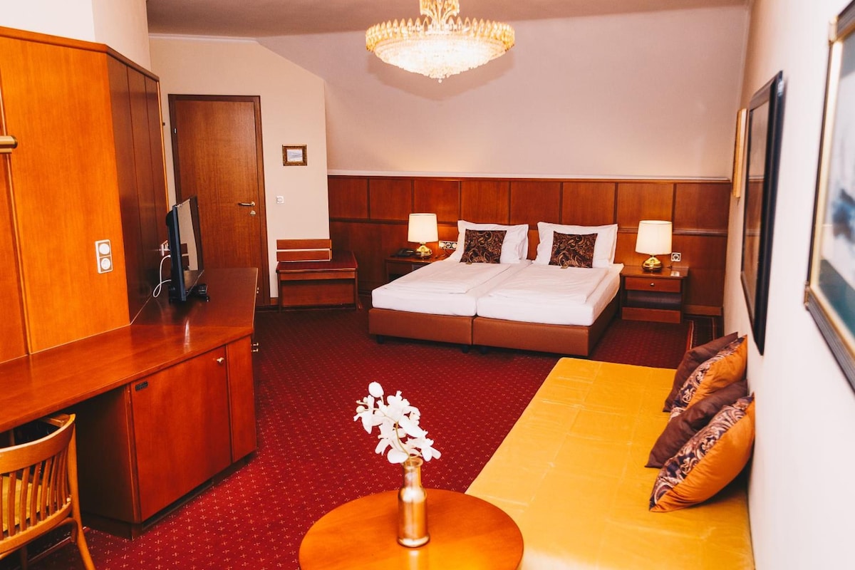 Habitación de hotel con cama, escritorio, silla, sofá, mesita, jarrón con flores y candelabro.