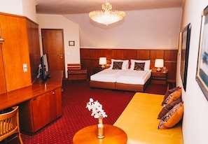 A hotel room with a bed, a desk, a chair, a sofa, a small table, a vase with flowers, and a chandelier. - Paul S Hotel (Knittelfeld)