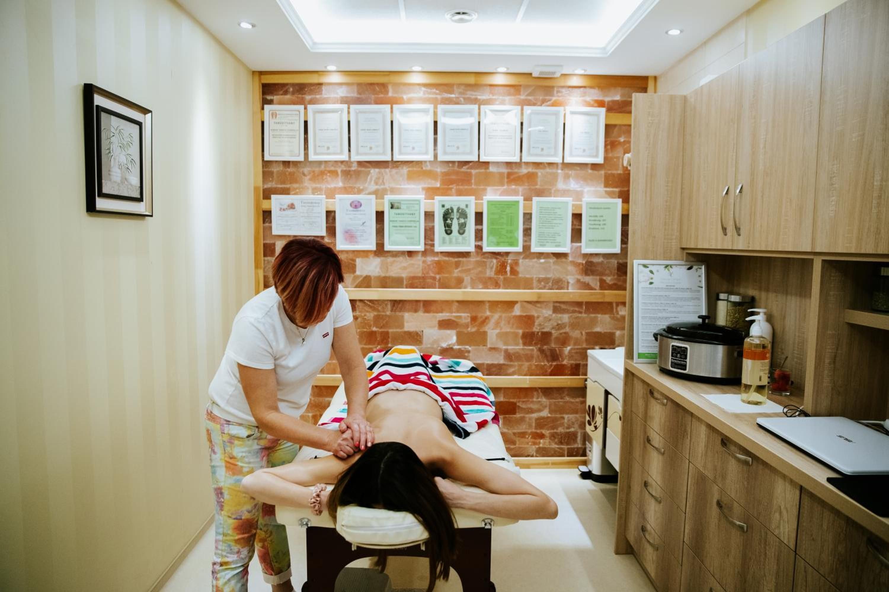 A massage therapy room with a client receiving a back massage, a wall with certificates, and a counter with a slow cooker and a laptop.