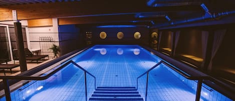 Una piscina interior con suelo de baldosas, una escalera central y un techo con vigas a la vista e iluminación.