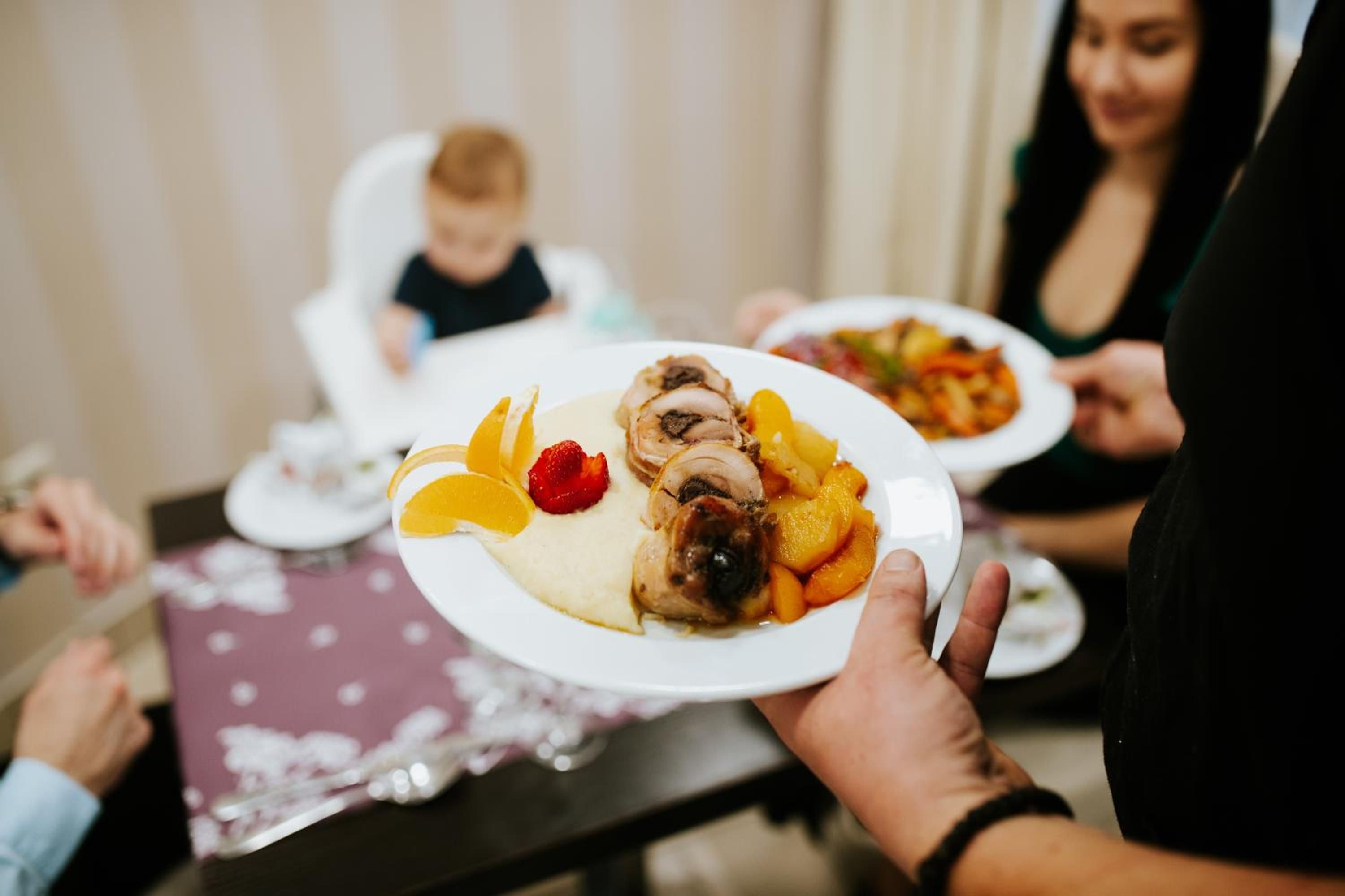 Eine Person hält einen Teller mit Essen, ein Kind sitzt in einer Hochstuhl und im Hintergrund ist eine weitere Person zu sehen.