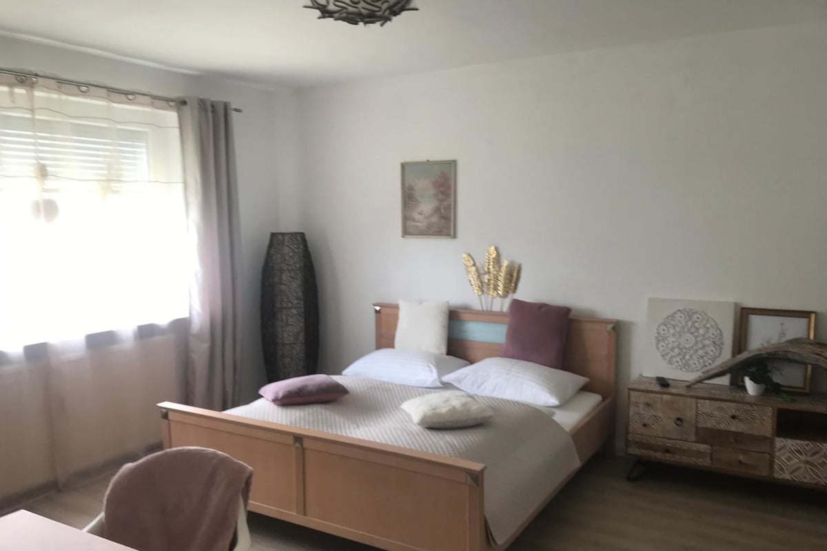 Un dormitorio con cama, una silla, una mesita de noche, un aparador y una ventana con cortinas.