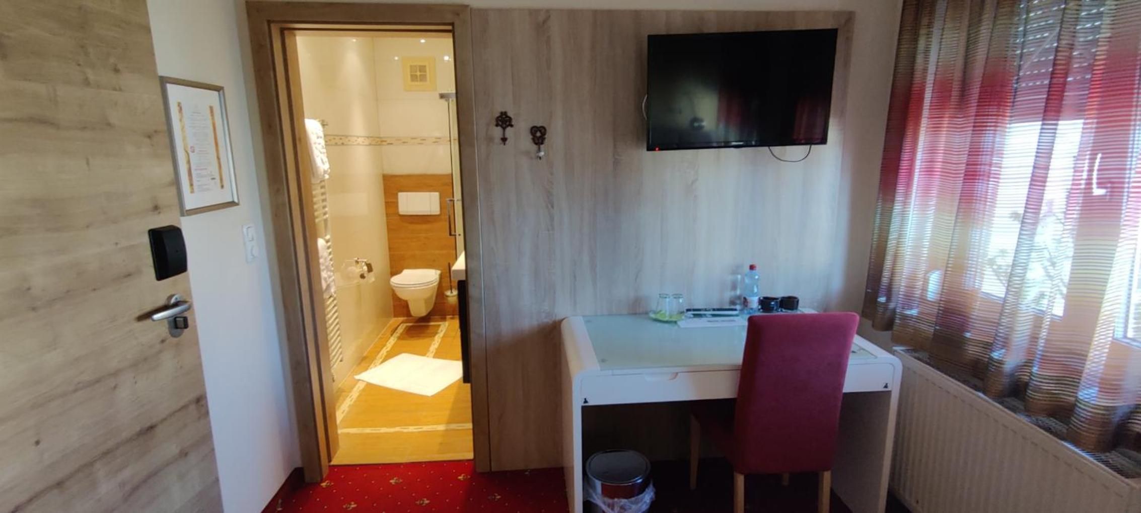 Ein Hotelzimmer mit Holzboden, Schreibtisch mit Stuhl, Wand-Fernseher und Fenster mit Jalousien.