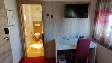 Ein Hotelzimmer mit Holzboden, Schreibtisch mit Stuhl, Wand-Fernseher und Fenster mit Jalousien.