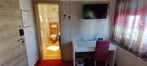 Habitación de hotel con piso de madera, un escritorio con silla, un televisor montado en la pared y una ventana con persianas.