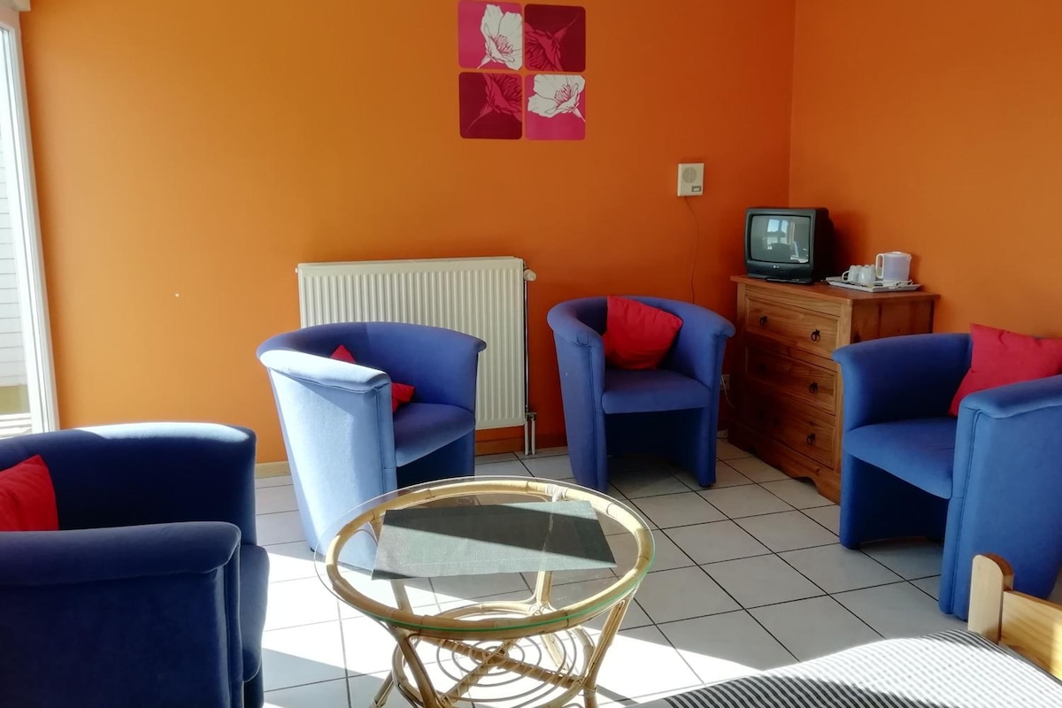 A room with two blue armchairs, a small glass table, a radiator, a television, and a wooden cabinet.