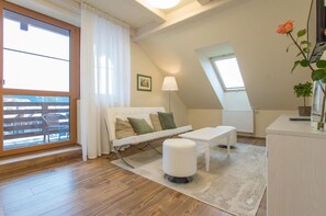 Una sala moderna con sofá, reposapiés y una mesa. Hay un balcón con vistas, una ventana con una planta y un cuadro en la pared.