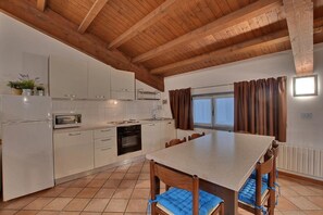 Una cocina con armarios blancos, techo de madera, mesa de comedor con sillas y una ventana con cortinas.