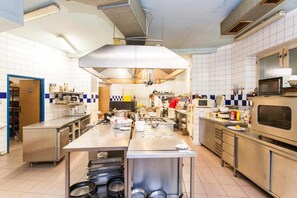 A commercial kitchen with stainless steel appliances, a large central island, and a person cooking in the background. - Hotel Grand (Tanvald)