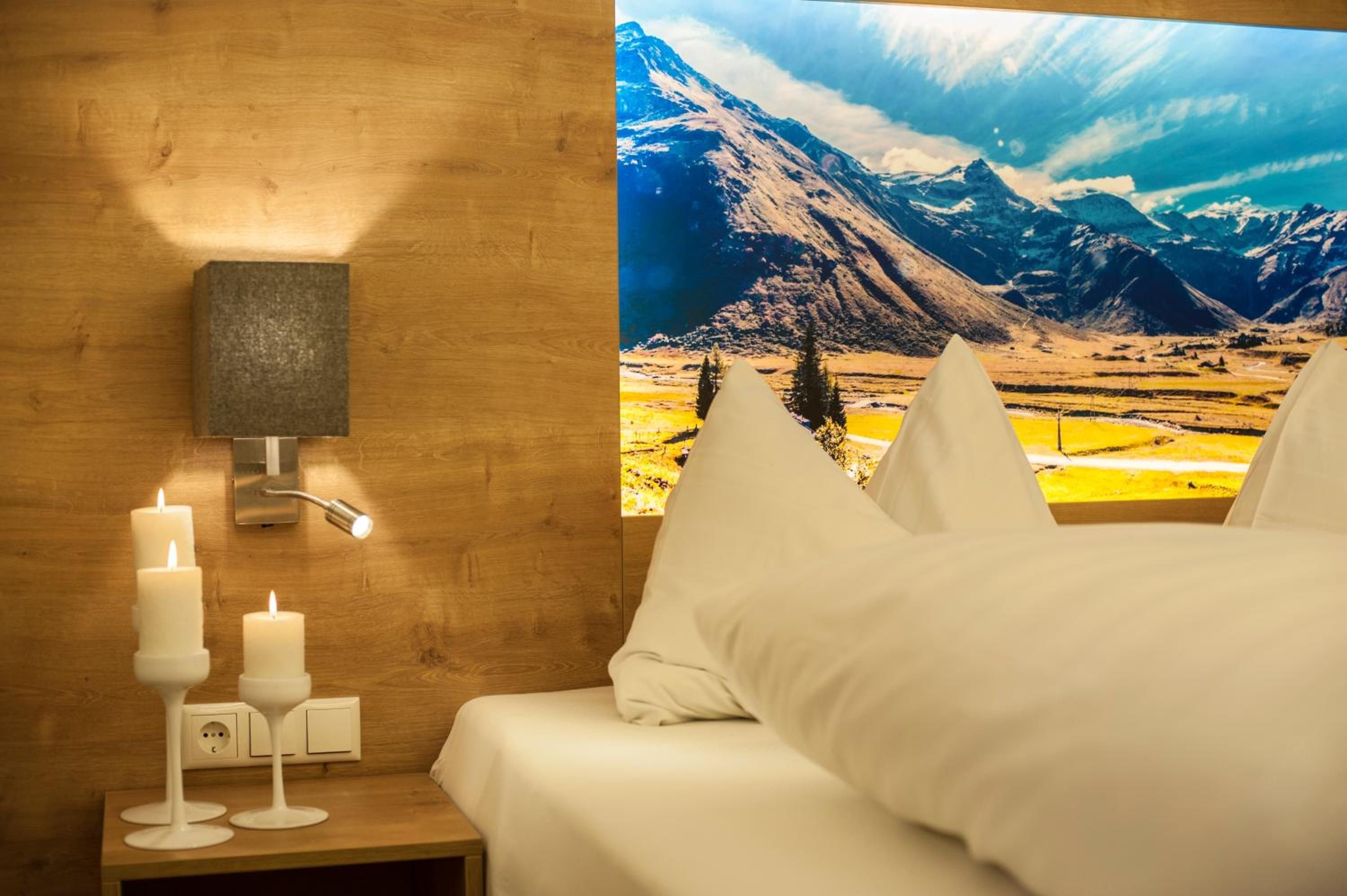 A hotel room with a bed, a nightstand with candles, a wall-mounted lamp, and a large window with a mountain view.