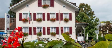 Un edificio de varios pisos con fachada simétrica, contraventanas rojas y macetas con flores en las ventanas.