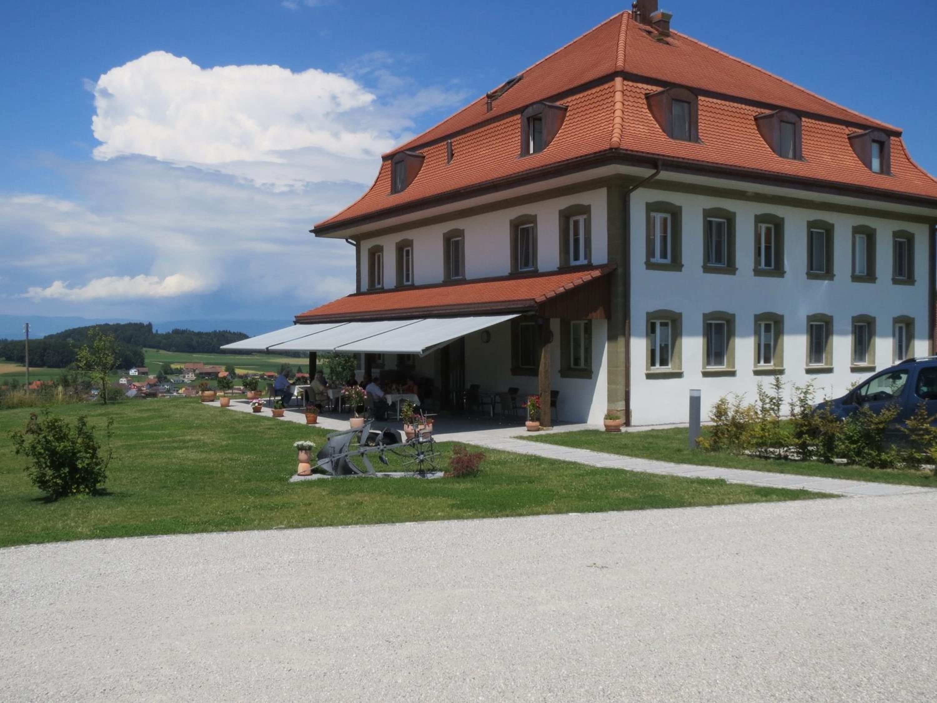 A two-story building with a red-tiled roof, a covered outdoor seating area, and a gravel driveway.