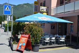 A street view with a pedestrian crossing sign, outdoor seating area with tables and chairs, and a large blue umbrella providing shade.