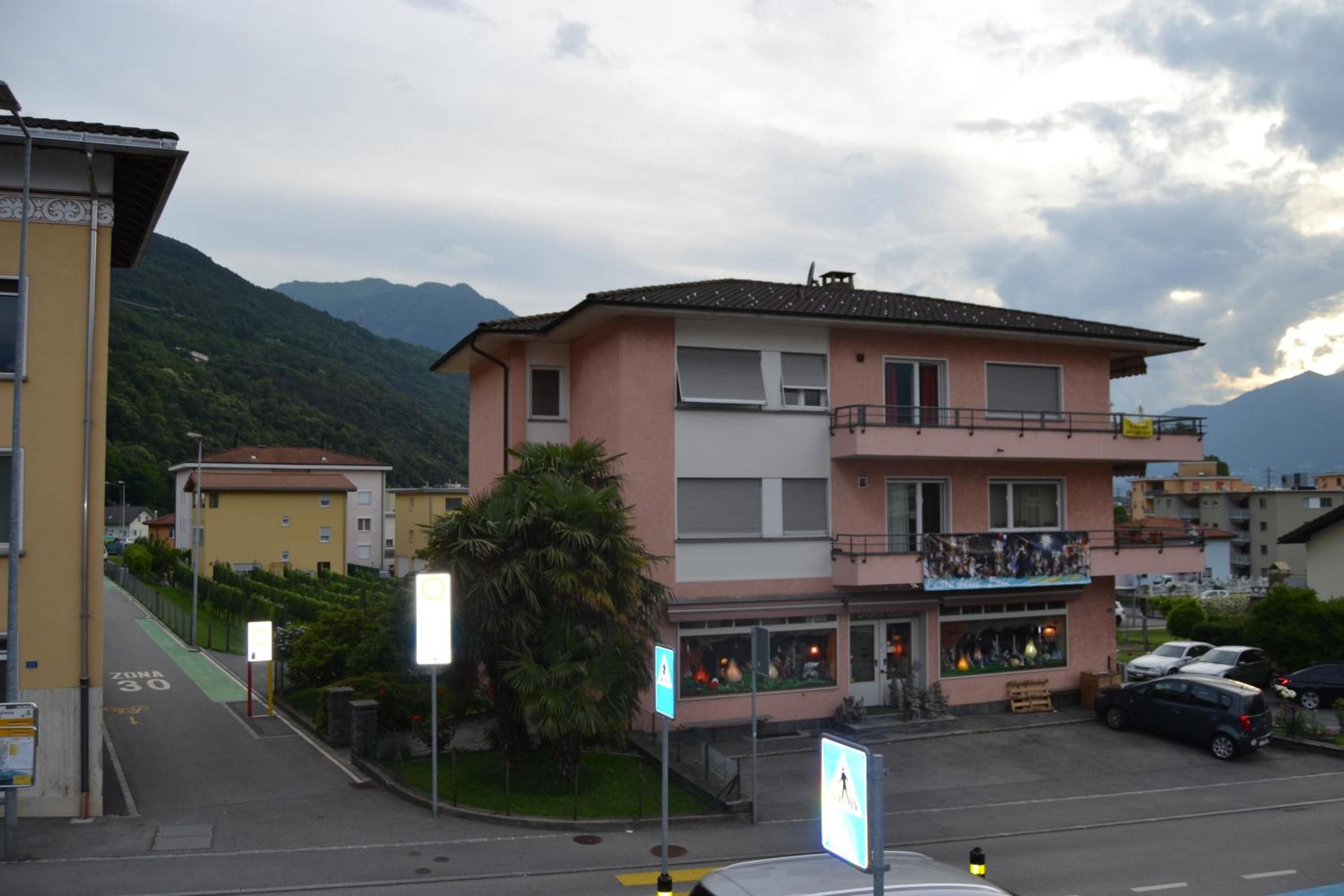 Un immeuble à plusieurs étages doté d’un balcon au deuxième étage, un espace vert avec un palmier, et une montagne en arrière-plan.