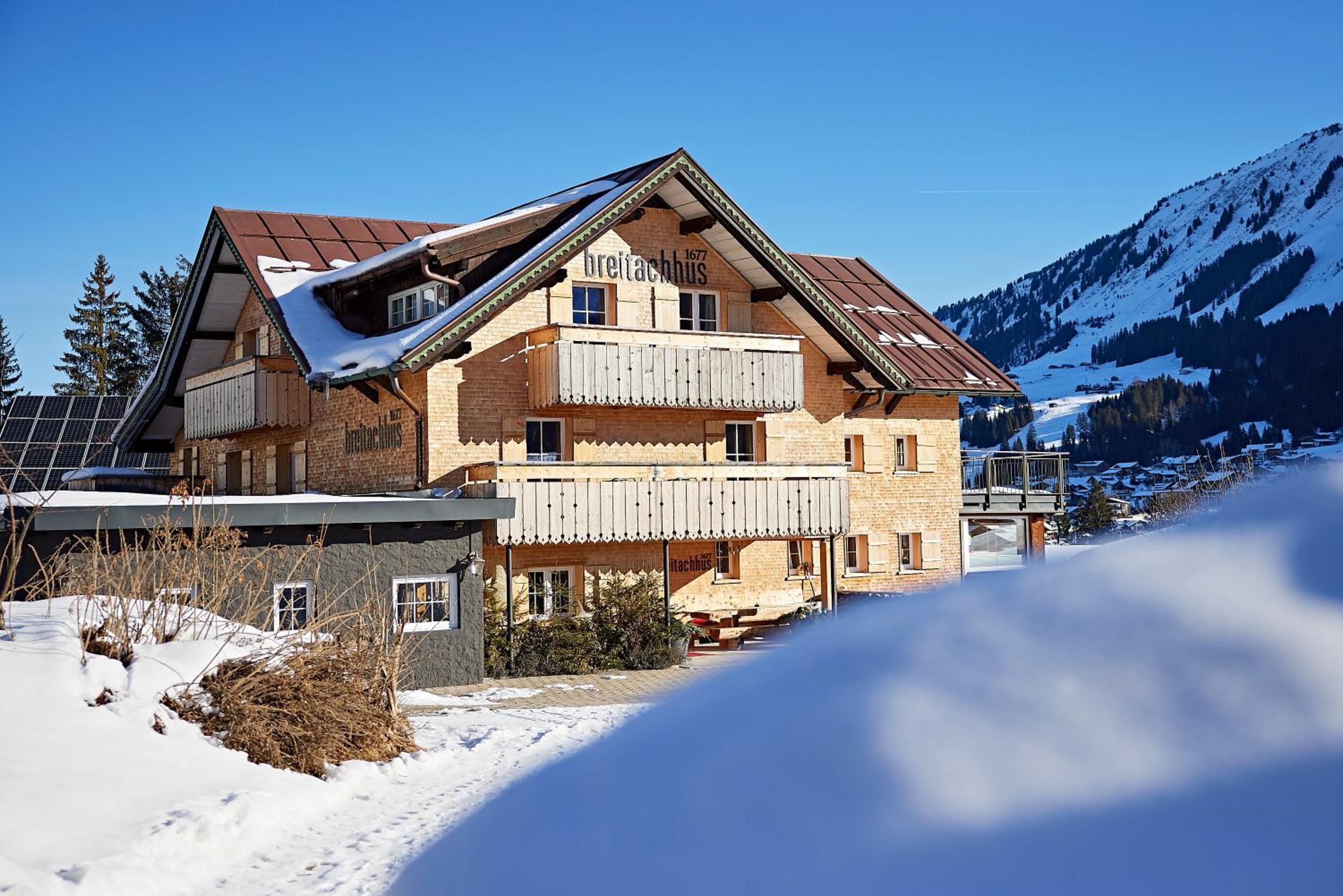 A two-story chalet with a wooden facade, a balcony, and a mountain backdrop.