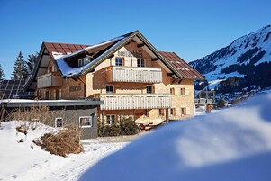 A two-story chalet with a wooden facade, a balcony, and a mountain backdrop.