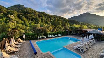 Piscina con sillas de descanso y sombrillas, rodeada de vegetación y montañas.