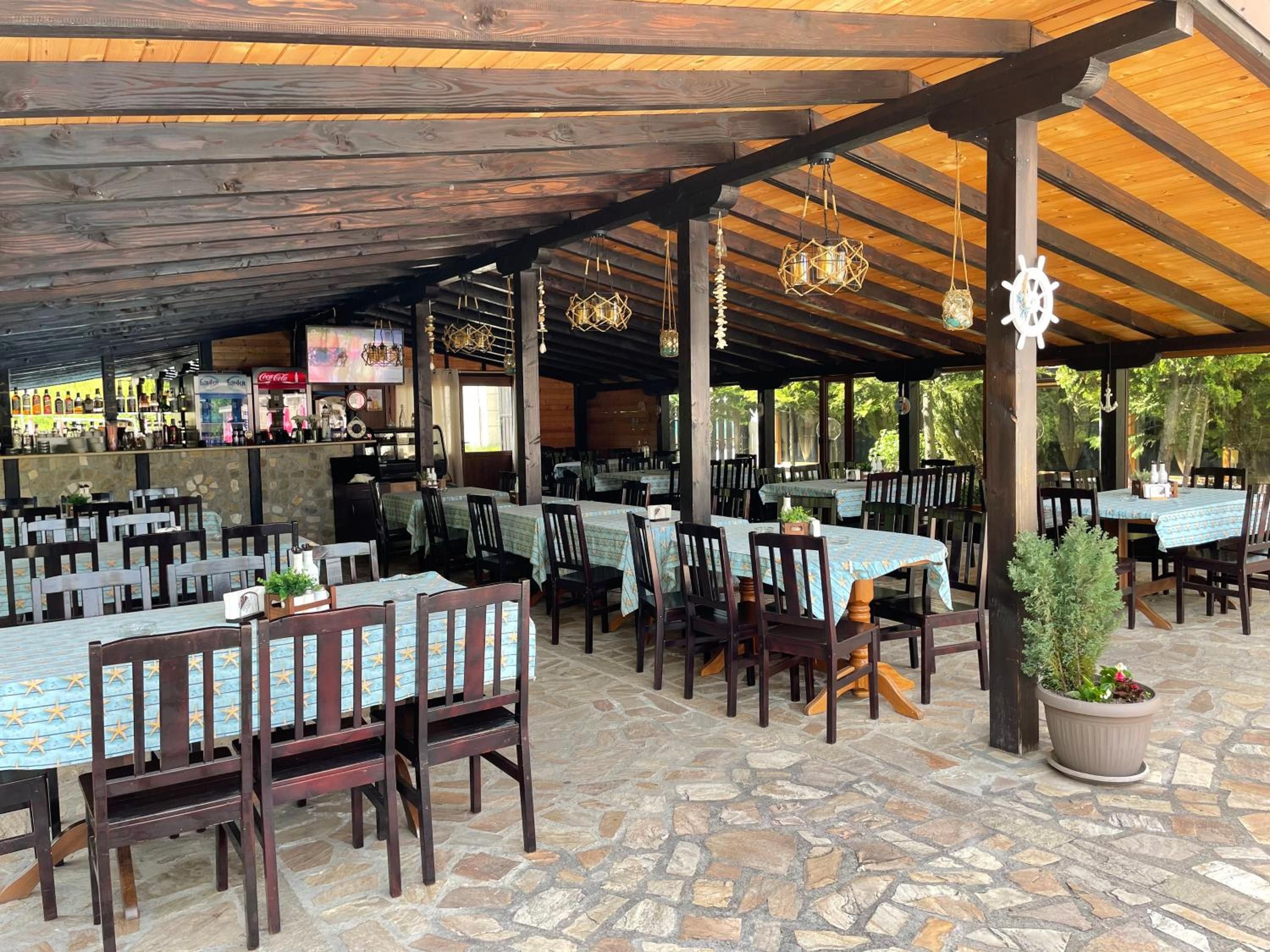 Un restaurante al aire libre con suelo de piedra, mesas y sillas de madera, y un área cubierta con techo de madera.