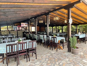 An outdoor restaurant with stone flooring, wooden tables and chairs, and a covered area with a wooden ceiling.
