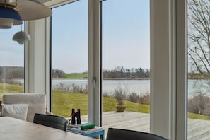 A room with a large window overlooking a lake, a table with binoculars, and a view of the outdoors. - Lovely holiday home by Vellerup Vig (Skibby)