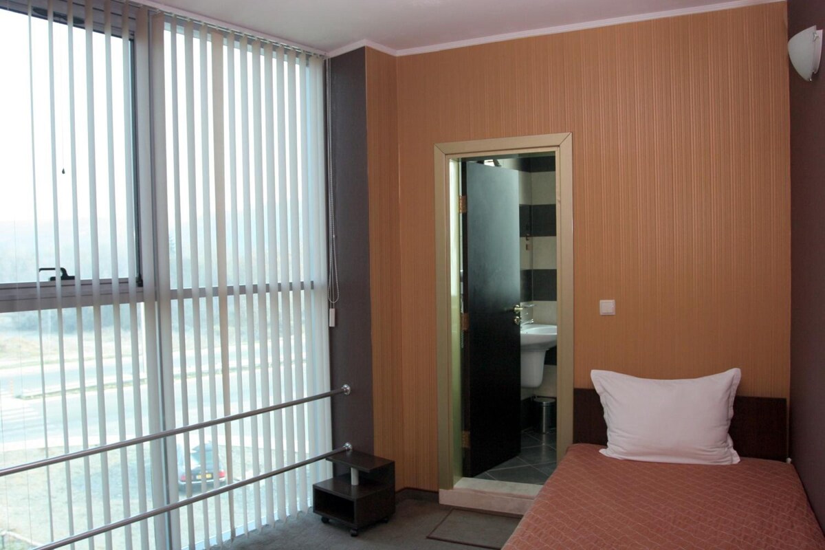 A hotel room with a bed, a bathroom visible through a glass door, and a view of a parking area.