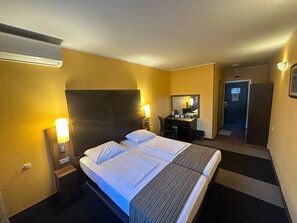 Habitación de hotel con cama, lámparas de noche, un escritorio con una silla, un espejo y una puerta.