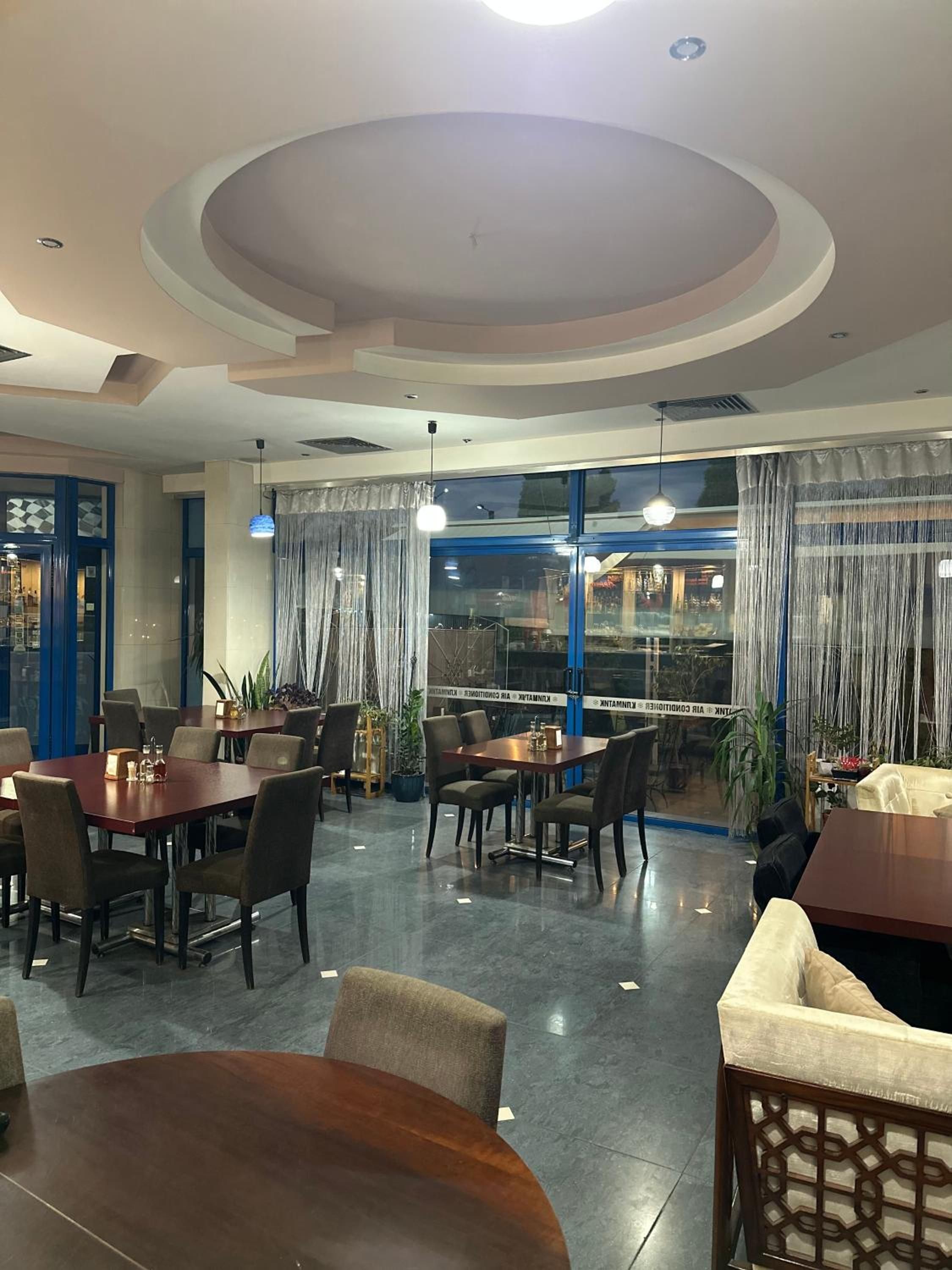 A restaurant with round tables and chairs, a view through large windows, and a modern ceiling design.