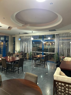 A restaurant with round tables and chairs, a view through large windows, and a modern ceiling design.