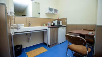 A compact kitchenette with a sink, microwave, and a small refrigerator. There is a chair and a table with a chair, and a trash bin is visible.